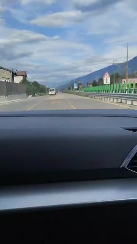 This video captures a view from the dashboard of a car driving on a road in Piani Selvetta, Italy. The scene is set during the daytime, under a sky with scattered clouds. The road appears to be a two-lane highway or a main road, with traffic moving in the same direction as the car.

In the foreground, the dark grey dashboard of the car is visible, taking up the bottom portion of the frame. The road ahead is clearly marked with yellow lines. On the right side of the road, a green metal barrier separates the road from what appears to be a hillside or embankment. Further along the road, there are streetlights and road signs. One prominent blue road sign with white text is visible, indicating directions to "SONDRIO" and "PIANI SELVETTA". There are also some buildings and trees visible on the left side of the road. In the distance, mountains form the backdrop, suggesting a mountainous or hilly region.

The activity is clearly driving. The mood is neutral, with no strong emotional cues evident from the static image. The weather appears to be pleasant, with a mix of sun and clouds. The time of day is likely late morning or afternoon, given the lighting conditions. The specific location cues from the road sign help to place the scene in Piani Selvetta, Italy.