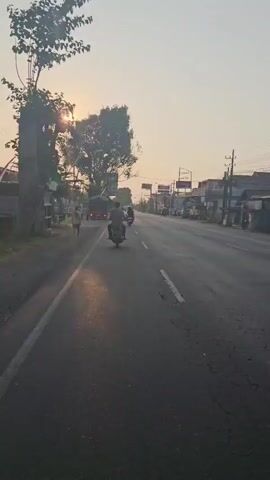 A motorcyclist rides on a road at sunset. The sun is visible through the trees on the left side of the road, casting long shadows. Other vehicles, including a truck and more motorcycles, are also on the road. The road appears to be a commercial area, with shops and buildings lining the right side. The sky is hazy with a pale orange glow.