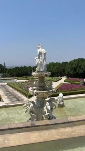The video shows a white marble fountain in a formal garden on a clear, sunny day. The fountain is multi-tiered and features sculptures of horses and figures.  Water spouts from the top tier into the tiers below.  In the background, there are geometrically designed flowerbeds separated by paved paths, with trees and possibly hills or mountains visible in the distance. A person can be seen walking on a path in the background. The camera slowly zooms in on the top portion of the fountain.