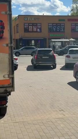 The video shows a parking lot in front of a commercial building on a sunny day. The building is painted yellow and houses several businesses, including "FIX price" and "Вкусвилл" (Vkusvill). A variety of cars are parked in the lot, including a light blue vintage car that drives into a parking space. A man wearing a cap, dark shirt, and jeans walks through the parking lot towards the camera. The ground is paved with bricks. A truck partially obscures the view on the left side of the frame. There are some trees in the background.
