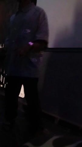 A person wearing a light-colored button-down shirt with the text "Buttlessin" on it and a baseball cap stands indoors, moving their hands slightly. Another person in a light blue t-shirt walks behind them. The scene is dimly lit, with a patterned wall to the left and a white wall to the right. There appears to be music playing in the background.