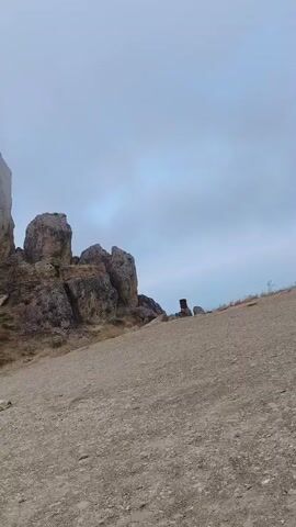 The video shows a rocky, mountainous landscape under a cloudy sky. The terrain in the foreground is a dirt and gravel slope. In the middle ground, large rock formations rise up, partially obscured by fog. To the left, a sheer rock face is visible. To the right, a tall, dark, cylindrical object stands upright. In the middle of the frame, on the dirt slope, a white dog is lying down. The camera appears to be moving slightly, panning across the scene.