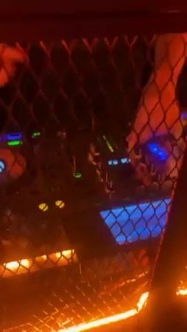 A man with a beard and long hair tied in a bun is operating a DJ mixer behind a cage. The mixer is lit with various colors, including blue and green. There are metallic silver balloons spelling out "PIRATA" behind him. The lighting is dim and red.