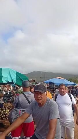 The video shows a crowded outdoor market scene. In the foreground, a person wearing a purple helmet and a purple garment is partially visible, facing away from the camera. A group of people are walking through the market, some appearing to be vendors and others shoppers. Tents and stalls are set up, displaying various goods, possibly clothing and other items. In the background, mountains are visible under a cloudy sky. The ground is dirt. The overall atmosphere suggests a bustling marketplace.