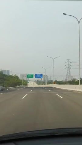 The video shows a forward-facing view from inside a vehicle moving along a multi-lane highway. The sky is overcast, and the environment appears to be urban or suburban, with buildings and power lines visible in the distance. Road signs are prominent, indicating directions to "Pagedangan Cisauk Karawaci" on a green sign and "Serpong Bintaro Pondok Indah" on a blue sign. The highway itself is paved, with clear lane markings, and appears to be relatively new or well-maintained, with construction barriers or temporary markings visible in some sections. The overall impression is of steady travel on a highway with clear directional signage.