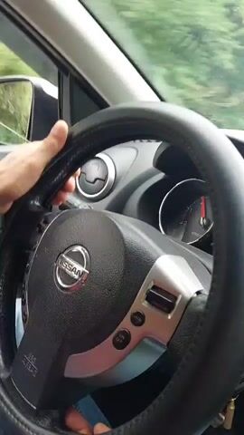 A first-person perspective video shows a person's hands on the steering wheel of a Nissan car. The steering wheel is black with silver accents. The dashboard is visible, including an air vent and a speedometer. The car appears to be moving, as evidenced by the motion blur of the scenery outside the window and the slightly blurred hands on the steering wheel. The driver is wearing blue jeans. The overall scene suggests a drive through a wooded or green area.
