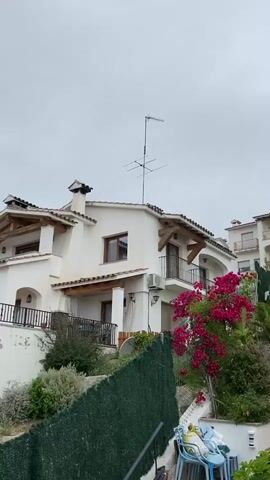 The video features a panning shot across a residential area in Vilassar De Dalt, Spain, under an overcast sky during the daytime. The camera moves from right to left, showcasing several houses built on a hillside.

The houses are predominantly white with terracotta roof tiles, and many feature wooden beams and balconies, characteristic of Mediterranean architecture. Antennas are visible on some rooftops. In the foreground, a vibrant bougainvillea bush with pink flowers adds a splash of color next to a green mesh fence. Further left, a swimming pool surrounded by artificial turf and outdoor lounge furniture is visible, suggesting a recreational area. Beyond the houses, hills covered with trees can be seen in the distance. No people or visible text are present in the video.