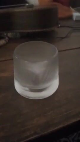 A short video shows a frosted glass cylinder on a dark brown wooden table. The glass appears thick and clear at the bottom, fading to a frosted finish as it rises. The table has visible wood grain and scratches. In the background, a dark object is partially visible, along with a couple of cables. The lighting is ambient and indoor, without any significant movements. The camera is mostly still. There is speech audible in the background.