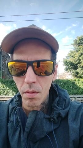 A selfie video shows a man from the chest up, looking directly at the camera. He is wearing a dark grey or blue hooded jacket, sunglasses with yellow-orange mirrored lenses, and a brown baseball cap. His expression is neutral with a slight hint of a smile.

The video begins with a background of green foliage, a blue sky with scattered clouds, and what appears to be a railway bridge or overhead structure. As the video continues, a large blue object moves behind the man. This object is a train or a similar vehicle, with the word "ARGENTINA" visible (and reversed due to reflection) on its side. The distinct sound of a train passing by is audible throughout the clip. The scene is set outdoors during daylight hours. The reflections in the man's sunglasses capture elements of the passing train and the surroundings.