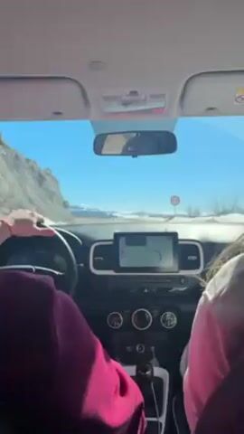 The video, shot from the driver's seat, shows a car navigating a road with a scenic backdrop. The driver, wearing a purple shirt, is visible on the left side of the frame. A passenger in a pink shirt sits next to them. The dashboard features a GPS navigation system guiding the route. Through the windshield, a clear blue sky frames a landscape featuring a mountain range covered in snow. The rearview mirror shows a reflection of "The North Face" logo. The road curves along a hillside, with some patches of snow visible on the side of the road. Music plays in the background, although this audio detail is separate from the visual description.