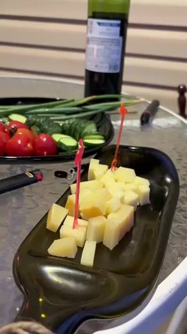A wine bottle, a plate of vegetables, and a dish of cheese are on a table. The vegetables include cherry tomatoes, sliced cucumbers, and green onions. The cheese is cubed and served with small red plastic skewers. A lighter, a jar, a candle, and a glass of red wine are also visible on the table. The setting appears to be outdoors with a striped wall in the background.