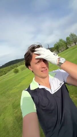 A man is standing on a golf course. He is wearing a white golf glove, a watch, and a golf shirt with a black vest over it. He touches his hair and smiles. He is holding a phone, presumably filming a selfie video.  The video then shows a golf cart with a golf bag on it. A hand wearing a white golf glove places a blue baseball cap on top of the golf clubs. The golf bag contains various golf clubs with green and red club covers, a white towel with red stripes, and a red pillow that says "Spalding" on it. The background is a green golf course with trees and a few buildings in the distance. The weather appears to be sunny.