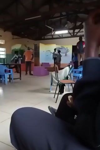 The video shows a person sitting in a chair, looking at a room where a play is being rehearsed. The room has a stage with a backdrop painted with ocean scenes. Several people are on the stage, seemingly practicing lines and movements. An adult man in an orange shirt stands on the side, possibly directing the scene. Several chairs are scattered around the room, some blue, some covered with blankets. The room has a high ceiling with exposed beams and fluorescent lighting. The person filming appears to be sitting in the audience area, watching the rehearsal.