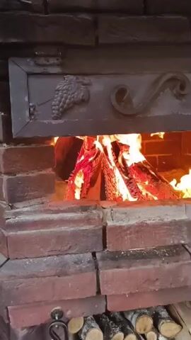 The video shows a close-up shot of a brick fireplace with a fire burning inside. The fire is made of logs, and flames are visible. Above the opening of the fireplace, there is a decorative metal plate with designs of grapes and a swirl. The bricks of the fireplace are red and appear to be old. At the bottom of the frame, there are stacks of cut firewood. The camera is positioned slightly below eye level and captures the scene from a static viewpoint.