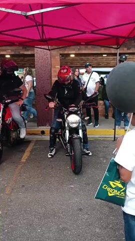 The video shows a motorcyclist sitting on a motorcycle, under a red canopy. The rider wears a red helmet, black jacket, jeans, and white sneakers. The motorcycle is black with red accents on the wheels. The rider appears to be posing for the camera. The environment includes a brick building, other motorcycles, and several people watching or walking by. The ground is asphalt, and a person carrying a "Chronus" lubricant shopping bag is partially visible in the foreground. The atmosphere seems to be casual, possibly at a motorcycle event or gathering.
