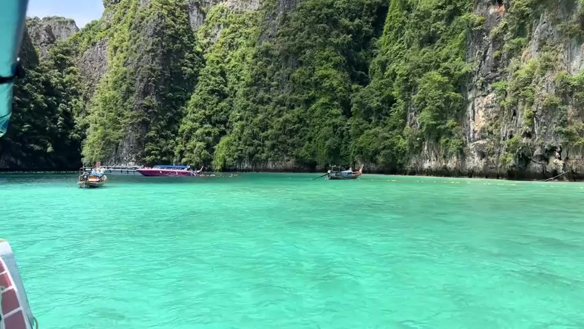 The video shows a view from a boat on a bright, sunny day. The water is a vibrant turquoise color and appears calm. In the background, there are large, lush green cliffs that rise steeply from the water. Several longtail boats are visible in the turquoise water, some of which appear to be anchored. A few people are in the water, swimming or wading near one of the boats. The overall impression is a tropical paradise scene.