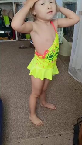 A young girl with blonde hair and blue eyes stands in what appears to be a room, possibly a home. She is wearing a neon yellow swimsuit with a ruffled skirt and an avocado print. The girl is touching the strap of her swimsuit with her hands. She stands on a carpeted floor. In the background, there are shelves with various items on them, and a curtain hangs on the right side of the frame.