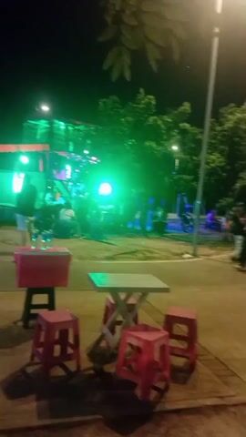 The video shows an outdoor scene at night. There are trees in the background and a bright street light illuminating the area. In the foreground, there are red stools and a small table. Further back, there are people walking and standing around. There are also some motorcycles parked along a road. A brightly lit structure with blue lights is visible on the left side of the frame. The scene appears to be a public space, possibly a park or a street with outdoor seating and entertainment.