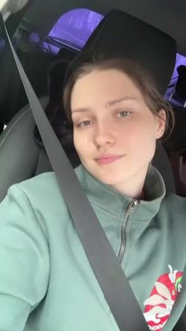 A young woman is recording herself in a car. She is wearing a light green zip-up sweatshirt, and the car’s seatbelt is buckled across her chest. She is smiling slightly and looking at the camera. The car appears to be moving based on the blurred view through the window. There is a headrest behind her and some items on the back seat. The interior of the car is visible, including the dashboard and part of the steering wheel. The lighting is natural.