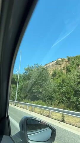 The video captures a drive through a scenic rural landscape in Greece, specifically within the Municipal Unit of Promachonas. The viewpoint is from inside a moving vehicle, looking out through the side window. The foreground features a car's side mirror and a portion of the car's interior frame.

The main subjects are the natural elements of the scene: a bright blue sky with a faint contrail, a steep, rocky hillside partially covered in lush green vegetation, and several trees. Dominating the middle ground is a tall white wind turbine, its blades slowly rotating, indicating it is actively generating power. The road appears to be a paved highway or main road, bordered by a silver guardrail and some scattered greenery.

The activity taking place is travel by car. The time of day is clearly daytime, likely late morning or early afternoon, given the bright sunlight and clear sky. The weather appears to be fair and sunny, typical of a warm day. The rocky hillside and the presence of wind turbines suggest a location that is either mountainous or hilly, and potentially exposed to consistent winds. There are no people visible in the media.

No text is visible in the video. The overall mood is one of peaceful travel and appreciation of the natural scenery.