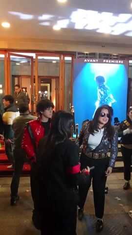 The video captures a bustling event, likely a film premiere, at the entrance of a building in Providencia, Chile. A red carpet is laid out, with numerous people gathered around. Several individuals are seen wearing attire reminiscent of Michael Jackson; one young man is in a glossy red jacket, and another person wears a sequined top, sunglasses, and a sparkling belt. Photographers and videographers are actively documenting the event with their cameras. In the background, a large screen displays an image of Michael Jackson with the prominent text "AVANT PREMIERE." Above the entrance, a sign is visible with the text "3 DE ABRIL SOLO EN CINEMAS" (April 3rd, Only in Cinemas), indicating a release date. The scene is brightly lit by overhead lights, suggesting an evening event. Attendees are seen walking, posing, and interacting, contributing to an excited atmosphere.