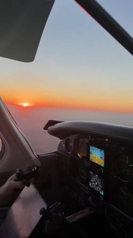 The video shows the interior of an aircraft cockpit during sunset. The sun is low on the horizon, casting a warm orange glow across the sky. Clouds are visible below the aircraft, appearing as a soft, hazy layer. The cockpit instruments are illuminated, with a pilot's hand visible on the yoke, controlling the aircraft. The overall scene conveys a sense of peaceful flight as the day comes to a close.