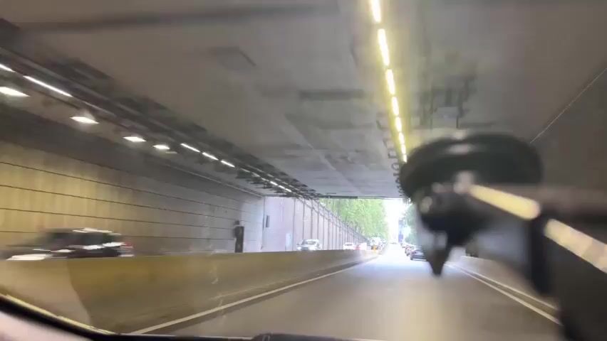 A first-person perspective video captures a drive through a tunnel and emerging onto a city street. The video begins inside a tunnel with fluorescent lights overhead and concrete walls. Traffic is visible ahead. The vehicle exits the tunnel and the scene transitions to an overcast day on a paved road lined with trees and a brick wall on the left. Several cars and a white van are on the road. The camera is mounted on the dashboard, with a suction cup mount visible in the foreground. The driving appears normal, with traffic moving steadily. The focus shifts between the road ahead and the foreground mount.