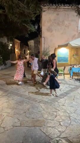 A group of people are dancing in a cobblestone courtyard at night. The group consists of adults and children. They are holding hands and dancing in a circle. A large tree with dark green leaves is in the background, casting shadows. There are buildings with illuminated windows behind the tree and to the right of the group. A table with chairs is visible to the far right, set up for outdoor dining. The lighting appears to be from lamps and ambient light from the buildings. The people are dressed in casual summer clothing.