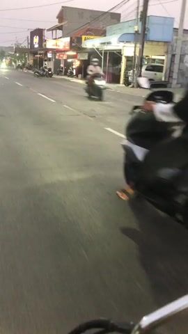 The video shows a street scene from the perspective of someone riding a motorcycle. The rider is behind another motorcycle with two people on it, a driver and a passenger, both wearing helmets. The street appears to be lined with shops or businesses. The time of day seems to be dusk or early evening, as indicated by the lighting conditions and the illuminated signs. The street is paved, and there are white lines marking the lanes. The overall atmosphere is that of a typical urban or suburban street scene during the evening commute or leisure time.