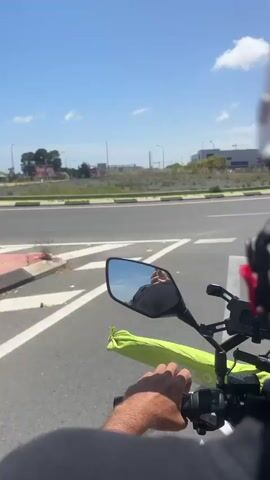 A first-person perspective video shows a person riding a motorcycle on a paved road under a bright blue sky with scattered clouds. The rider's hands are visible on the handlebars, and a green bag is attached to the motorcycle. A mirror reflects the rider's helmet and upper body. The road has white lane markings and a green-painted bike lane along the side. In the background, there is a roundabout sign with blue and white arrows, streetlights, and buildings, suggesting an urban or suburban environment. A black car is driving on the road ahead and to the right of the motorcycle. The video captures the movement of the motorcycle as it navigates the road.