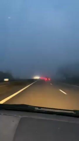 A car is driving on a highway on a foggy day. The car's headlights illuminate the road ahead. Several other vehicles are ahead, their headlights and taillights visible through the fog. The road is divided by solid white lines. There is a barrier on the left side of the road and trees visible through the fog on the right. The sky is a dull gray. The dashboard of the car in which the video is taken is visible at the bottom of the frame. Music is playing in the background.