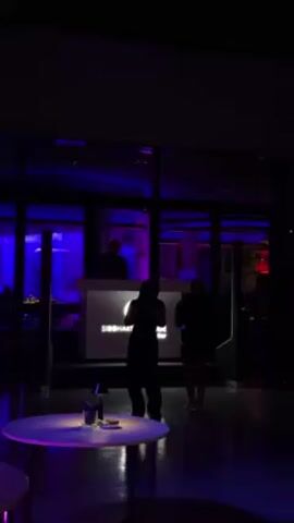 The interior of a darkly lit nightclub is shown. Two women in silhouette dance in front of a raised DJ booth. The booth is lit from behind, showing the DJ and the name “Siddharta Lounge”. Round tables stand in the foreground with candles and drinks on them. People walk through the frame. Blue and red lights fill the room. A mirrored window separates the club from the outside. The video appears to be taken on a phone.