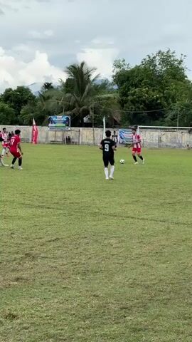 The video shows an outdoor soccer game on a cloudy day. In the foreground, a player in a black jersey with the number 9 on the back stands facing away from the camera, holding a soccer ball. To the left, three players in red jerseys are visible. One player is on the far left, facing right, another is in the center, facing the camera, and a third is to the right of the center player, facing left. In the mid-ground, another player in a black jersey is running toward the left, and a player in a red jersey is near the goal on the right. A fence with advertising banners runs along the back of the field, and beyond that, trees, buildings, and mountains are visible. The grass on the field appears somewhat worn and dry in places. The overall atmosphere suggests a casual or amateur sporting event.