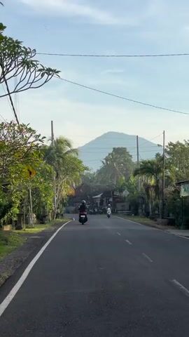 The video captures a road scene in a tropical setting, possibly early in the day. The camera faces down a black asphalt road with a white painted dividing line. Several motorbikes, each with a rider, are moving towards the camera along the road. Lush, green tropical vegetation borders both sides of the road, including palm trees. Power lines run across the sky above the road. In the distance, a large mountain can be seen against a blue sky with some clouds. The setting suggests a rural or semi-rural area in a warm climate.