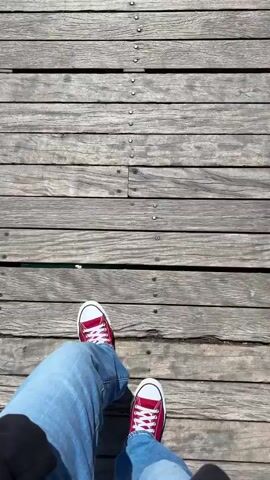 A person wearing jeans and red sneakers walks along a wooden pier towards the end. There are other people walking ahead on the pier as well.  On either side are benches and the railing. The pier extends out over the ocean. There is a row of lights hanging overhead. On the other side of the ocean is a town with buildings and a large hill in the background. There is a coin-operated telescope on the pier.