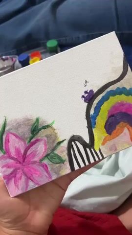 A hand holds a canvas painting with a colorful design. On the left side of the canvas, there is a pink flower with green leaves. On the right side, there is a black curved line with rainbow-colored stripes and black vertical lines. There are small bottles of paint in the background. The canvas is positioned on a person's lap covered with a blue fabric.