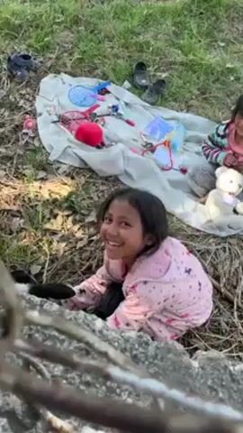Two young girls sit on the ground beside a blanket with various toys scattered across it. The ground is covered in grass and dry leaves. The girl in the foreground wears a pink pajama top with a star pattern and black pants. She is reaching for a small black and white lamb wearing a purple collar. The second girl sits on the blanket, partially obscured, wearing a striped shirt. A teddy bear sits next to her. In the background, pairs of sandals and shoes are scattered on the blanket. The video is shot from behind a concrete barrier.