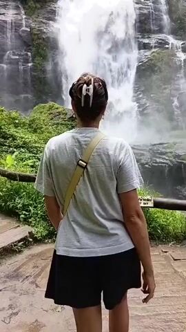 A young woman with her back to the camera walks towards a cascading waterfall. She is wearing a grey t-shirt, black shorts, and white sneakers with green accents. A light-colored crossbody bag is slung over her shoulder. She turns to face the camera, smiling, and poses in front of the waterfall. The waterfall is tall and powerful, surrounded by lush green vegetation and rocky cliffs. A wooden railing is visible in the foreground, and a sign reading "DO NOT SIT" is attached to it. The ground is wet and appears to be made of stone or concrete. The overall scene is a natural landscape with a person enjoying the view.