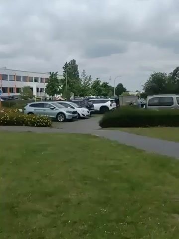A video shows a parking lot with several cars. The cars are parked in rows. There are also trees and bushes in the background. The sky is cloudy.