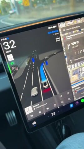 The video shows the interior of a Tesla car. The dashboard displays a self-driving interface with a map and a visualization of the car's surroundings. The car is on a highway with other vehicles visible. The driver's hands are on the steering wheel, and the car appears to be navigating a curve. The video is taken from the driver's perspective.