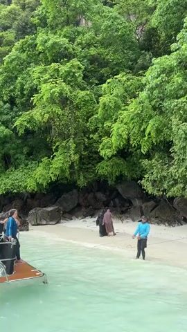 The video shows a tropical beach scene with lush green trees covering a rocky cliff face. The foreground features clear, turquoise water with some gentle waves. Several people are visible on the sandy shore and in the water.

On the beach, two individuals are walking away from the camera, one wearing a light brown headscarf and dress, and the other in a darker outfit. Further along the beach, a person in a bright blue jacket and dark pants is standing in the shallow water, holding a rope that extends towards the water. Another person in a white t-shirt and dark shorts is wading into the water. A person in a black t-shirt and shorts is also standing in the water. In the lower part of the frame, two individuals, one wearing a dark blue jacket, are partially visible in the water.

The scene appears to be a popular tourist spot, possibly a beach accessible by boat, as a portion of a boat is visible in the bottom left corner. The overcast lighting suggests it might be a cloudy day or early morning/late afternoon. The vibrant greenery and clear water are indicative of a tropical location like Ton Sai, Thailand. The people's attire and relaxed postures suggest a leisurely day at the beach, possibly a guided tour or excursion. There is no visible text in the video.