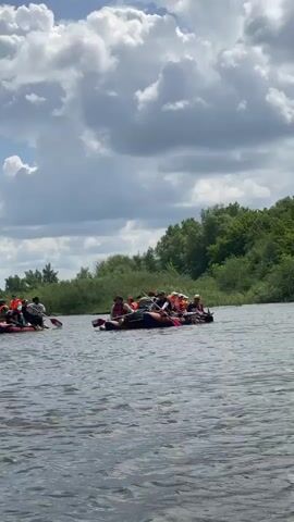 The video shows a wide shot of a body of water, likely a river, on a partly cloudy day. In the foreground and middle ground, several rafts or inflatable boats are visible. The raft closest to the camera is filled with people, most of whom are wearing life jackets. They appear to be paddling or navigating the water. In the background, there is a tree line and a sandy bank. Another raft with people is further to the left, also seemingly in motion. The overall impression is of a group activity on the water.