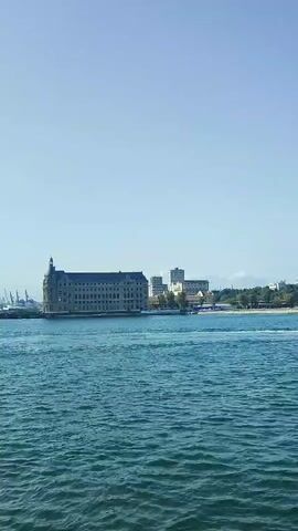 A wide shot video shows a large building on a body of water. The building is tan with many windows. There are some smaller buildings behind it, as well as some trees. A large blue ship is docked to the left of the building. In front of the building and to the left, a smaller white boat is moving across the water. The sky is a clear blue. The water is a deep blue-green with small waves.
