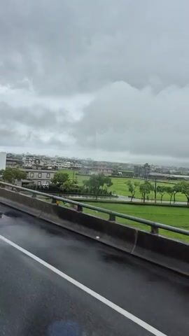 The video is a first-person perspective shot from a moving vehicle, likely a bus or train, showing a landscape under a cloudy sky. The foreground features a wet road with lane markings and a concrete barrier with a railing. Beyond the barrier, a rural landscape unfolds, characterized by green fields, scattered houses with tile roofs, and trees. The overall atmosphere is overcast and appears to be either rainy or recently rained upon, indicated by the wet road surface and the muted colors of the sky. The motion of the vehicle creates a slight blur, but the scene remains clear enough to discern the elements of the environment. No people are visible.