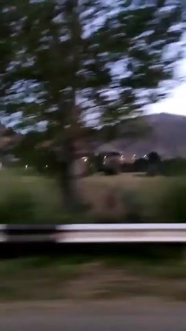 The video shows a blurry view from a moving vehicle. The scene appears to be rural, with mountains in the distance and trees in the foreground. There is a guardrail visible along the side of the road. The sky is a pale blue, suggesting it is either dawn or dusk. No people or specific actions are discernible due to the blurriness and speed of the vehicle.