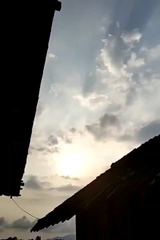 The video shows a view of the sky between two silhouetted structures. The sun is visible through scattered clouds, casting rays of light. The left structure is a dark, vertical shape, possibly a building or wall. The right structure is a dark, angled shape, resembling a roof. The sky is a mix of blue and grey, with fluffy white clouds. The overall scene suggests an outdoor setting, possibly during sunrise or sunset.