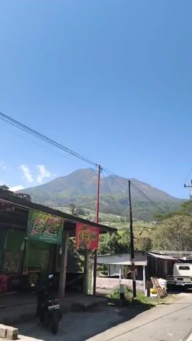 The video begins by showing a mountain peak against a clear blue sky. In the foreground, there is a road with power lines overhead. To the left, there is a building with a red sign that has yellow text. Further down the road, a white pickup truck is parked. The video pans to the right, revealing more of the mountain and a hillside covered in crops, possibly vegetables, leading down to buildings and trees. There are also more power lines and utility poles visible throughout the scene. The overall environment appears to be a rural or mountainous area with evidence of agriculture and local businesses.