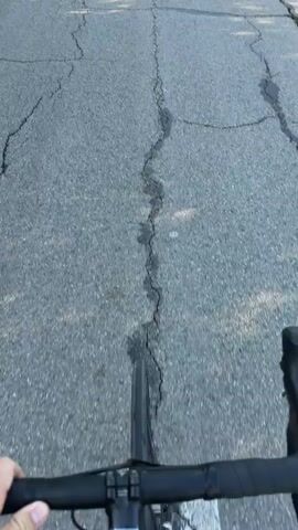 A first-person perspective video shows a person riding a bicycle on a paved road. The road is cracked and has some loose gravel. There are trees and bushes on both sides of the road. The sky is clear and blue. The video shows the handlebars of the bicycle, the rider's hand on the handlebars, and the road ahead. There is a sticker on the stem of the bicycle. The rider is moving forward at a moderate pace.