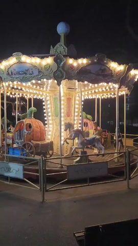 The video shows a small merry-go-round, illuminated with lights, rotating at night. The merry-go-round has horses and pumpkin-shaped carriages as its ride options. It is enclosed by a low, metal fence. The background is dark, suggesting a park or outdoor area at night. There is a bench in the lower right corner of the frame. The merry-go-round is stationary. There are no people riding it.