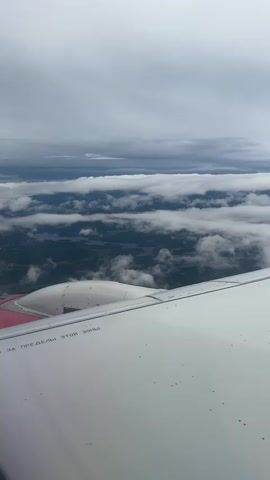 The video shows a view from an airplane window as it flies over a landscape. The wing of the airplane is visible in the foreground, taking up the bottom right portion of the frame. The wing is white with some black speckles. Text in Russian is visible on the wing, reading "ЗА ПРЕДЕЛЫ ЭТОЙ ЗОНЫ" which translates to "BEYOND THIS ZONE". In the middle ground, there is a forest canopy with bodies of water interspersed throughout. The background consists of layers of clouds. The sky is overcast, with varying shades of gray and white clouds. The overall impression is of a cloudy day seen from high altitude.